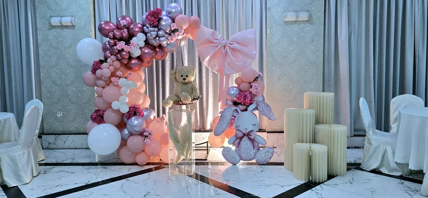 Blush balloon arch with a big bow, a teddy bear, a toy bunny and candles, set before rows of white chairs