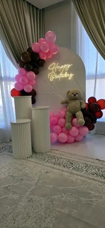 Pink balloons and a teddy bear at the foot of a cream arch with a gold Happy Birthday script